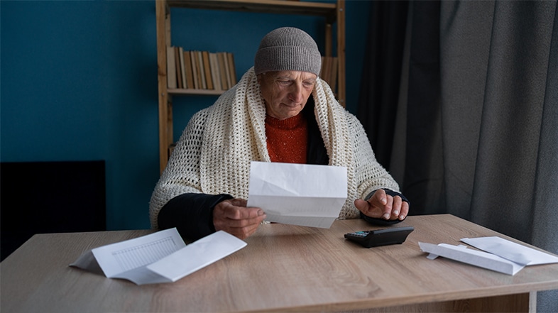 Older man wearing a knit cap and blanket sits at a wooden table reading a letter, with a calculator and scattered bills nearby, suggesting he is paying bills at home.